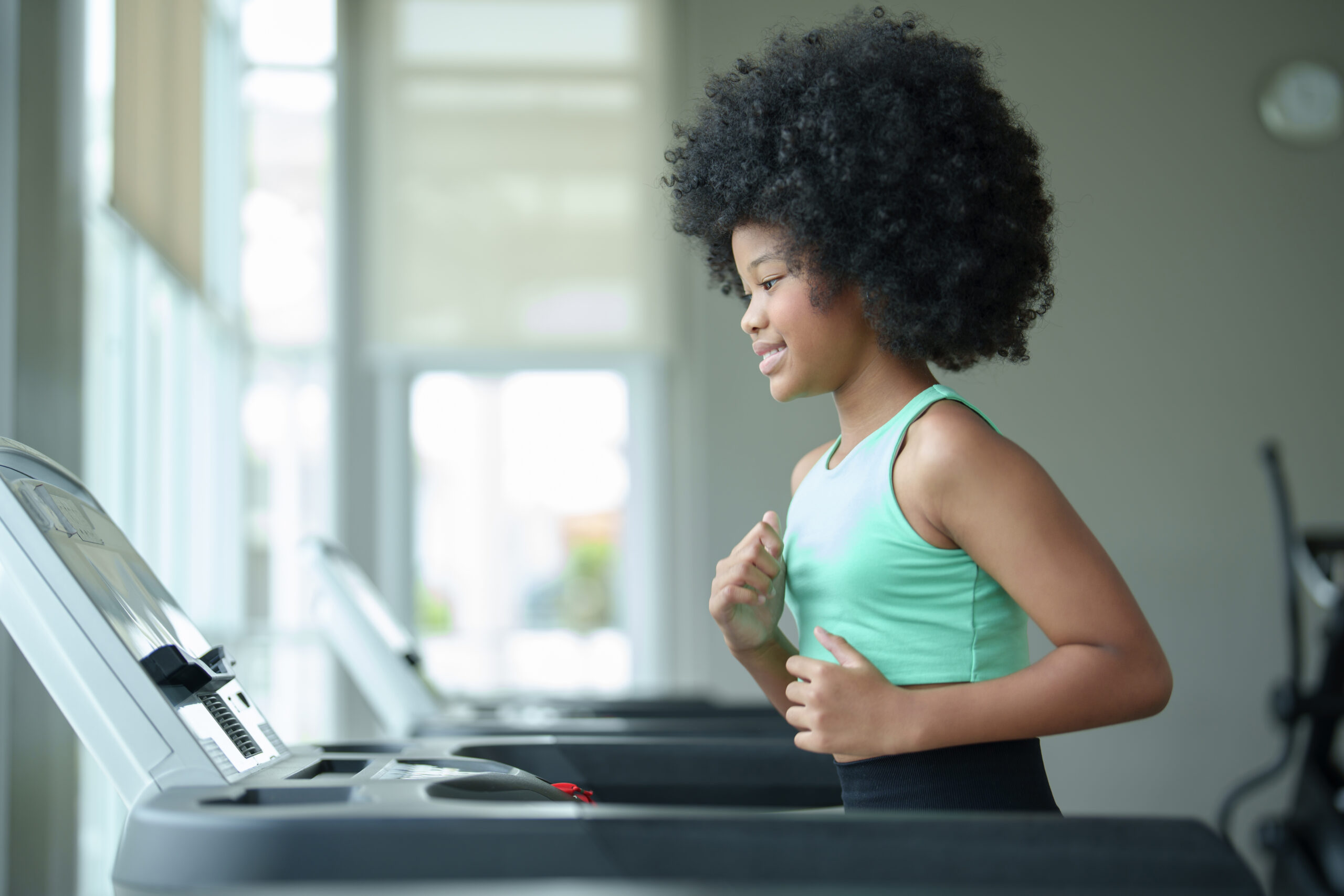 Young girl with curly hair exercises on treadmill in bright gym, wearing green tank top and black leggings, focused and determined, promoting a healthy lifestyle.
